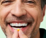 Smiling man holding a small yellow pill near his mouth.